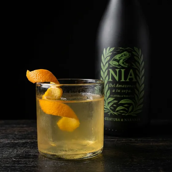 Vaso corto con piel de naranja, junto a la botella de Nía Guayusa & Naranja