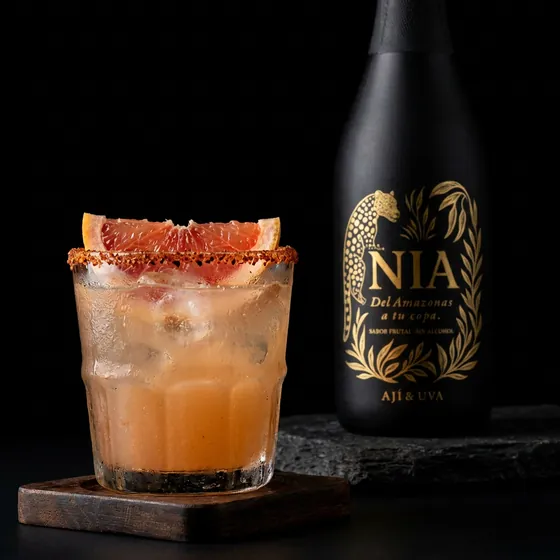 Vaso con borde escarchado y toronja, con la botella de Nía Ají Amazónico & Uva al fondo
